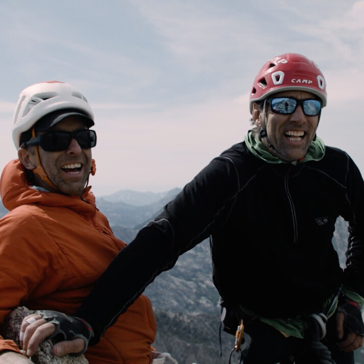 On the left stands Timmy O&rsquo;Neill wearing an orange jacket and helmet, smiling with sunglasses on. To his right stands blind climber Erik Weihenmayer in a black jacket and red helmet. Both men are on a mountain summit, appearing happy and enjoying the vast mountainous landscape in the background. The image captures the spirit of climbing, adventure, and determination.