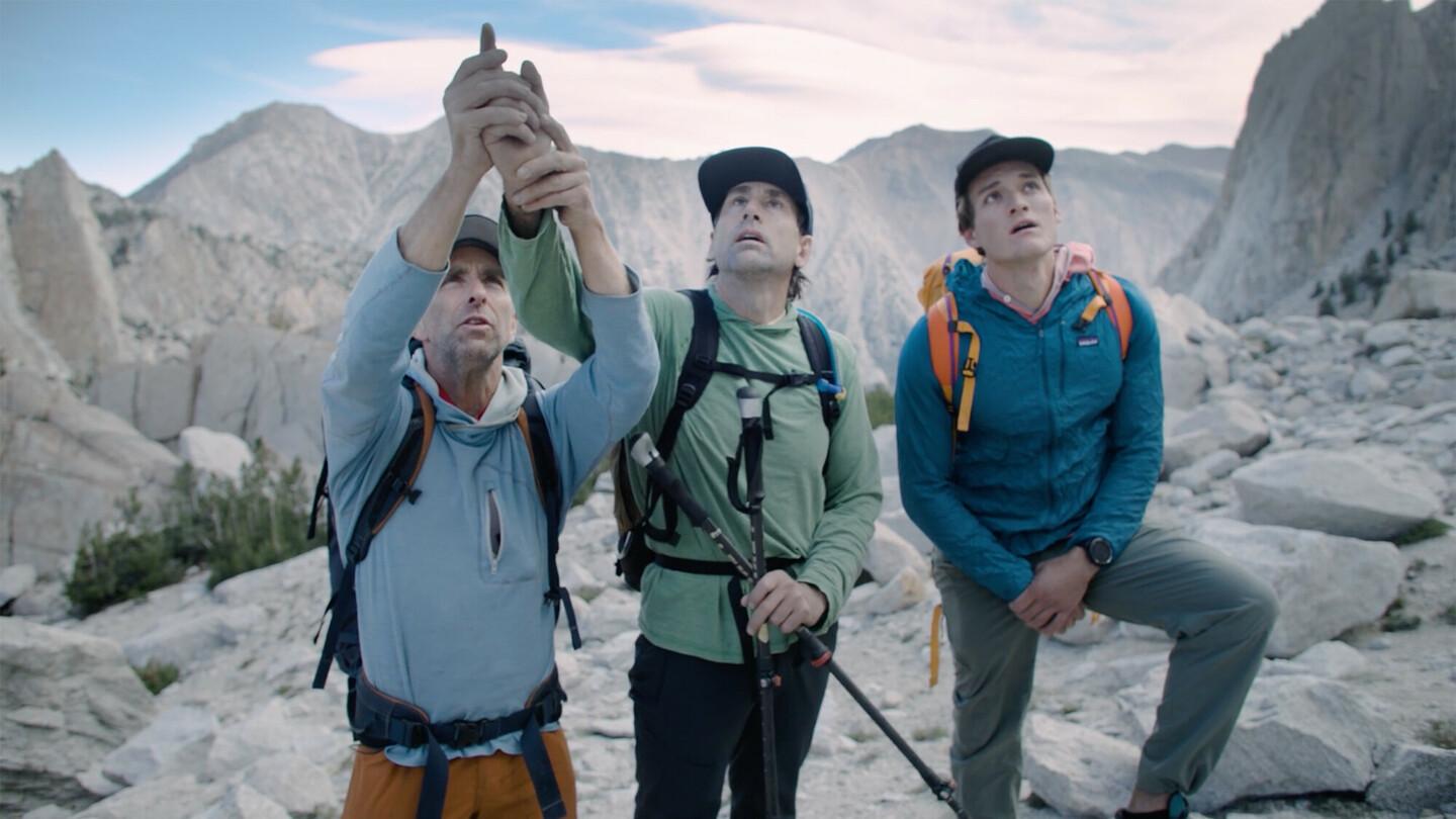 Three hikers in outdoor gear with backpacks stand in a rocky mountain landscape. The man on the left guides the blind man’s hand in the center to help him feel the climbing route and surrounding mountains.