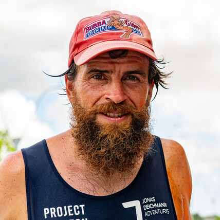 Portrait of Jonas Deichmann, a man with a thick beard and red cap featuring the Bubba Gump Shrimp Co. logo. He wears a dark tank top.